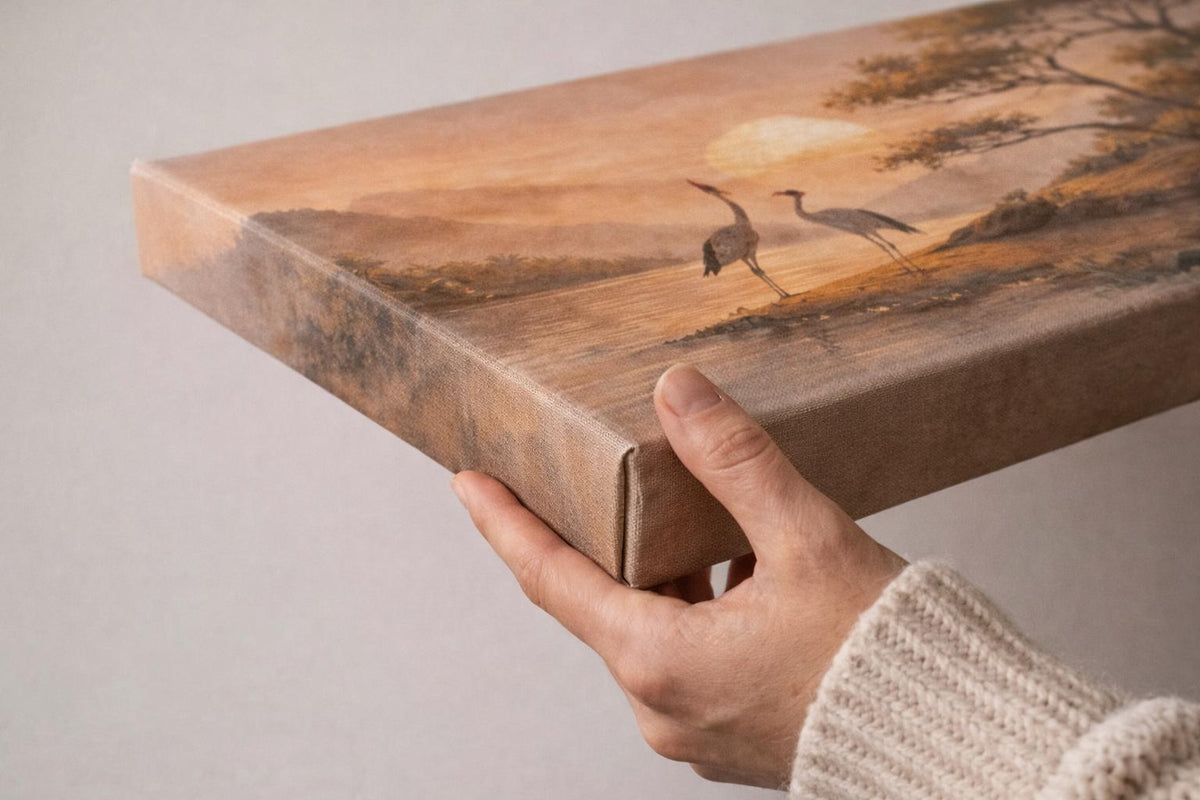 May include: A canvas print showing a tranquil landscape with two cranes by water at sunset. The artwork uses warm orange, brown, and beige tones. The canvas is held by a person wearing a beige sweater
