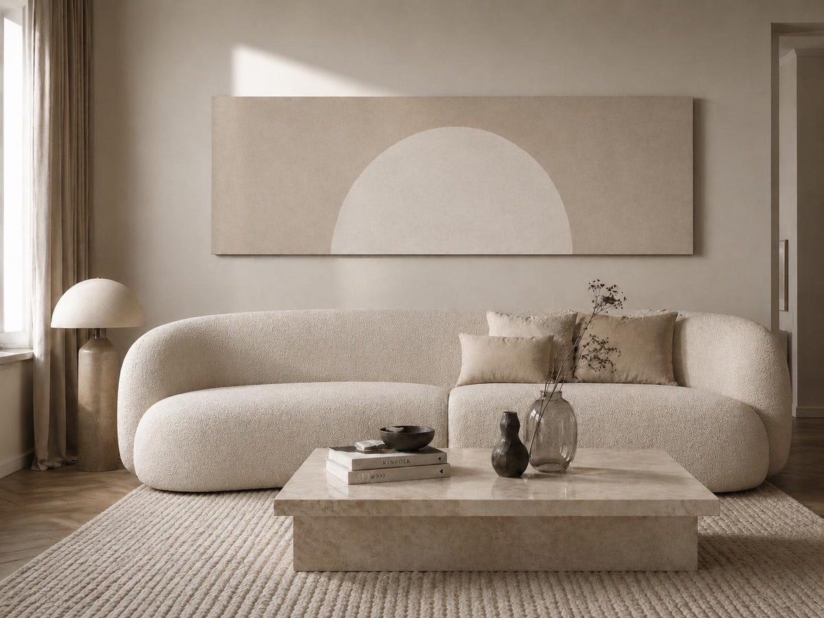 May include: A long, rectangular wall art piece featuring a large, pale beige semi-circle against a muted taupe background. The artwork is displayed above a curved, cream-colored textured sofa, a low stone coffee table, and a tall, rounded floor lamp.